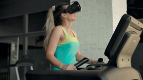 Beautiful caucasian woman in VR helmet walks on treadmill in gym. The woman is broadcasting the surroundings in the form of a park or forest path as VR content. Slow motion portrait.