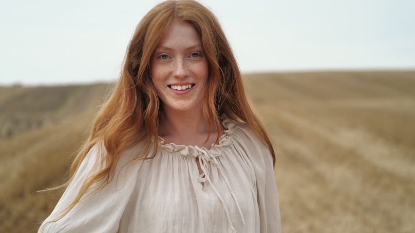 Ginger Woman  running on golden Field turning to the camera and smiling.  Woman with red hair and freckles touching her long Red Hair, walking on wheat meadow