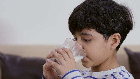 Close-up shot of a small adorable boy drinking a glass of milk at his home . A little boy in his living room finishing a glass of milk wiping his mouth with hands afterwards