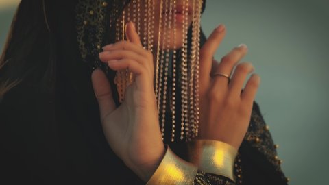 Portrait of young a beautiful oriental mysterious woman. The face is hides by golden veil. head is covered with a black scarf. Girl in traditional Egyptian Bedouin dress bracelets, accessories on hand
