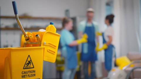 Focus on yellow bucket with cleaning tools and team of janitors giving high five on blurred background. Happy cleaning company personnel gesturing high five after successful clean