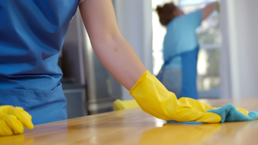 Close up of person in gloves and apron polishing or wiping wooden table with woman cleaning window on blurred background. Cleaning service team with professional equipment during work Royalty-Free Stock Footage #1059142703