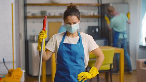Portrait of woman maid in apron gloves and mask positing at camera in apartment with colleagues cleaning on background. House cleaning service crew in protective mask working