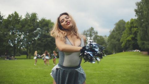Portrait of happy cheerleader woman dancing with pom-poms in park. Attractive young cheerleading lady in grey and white uniform performing cheering dance on field