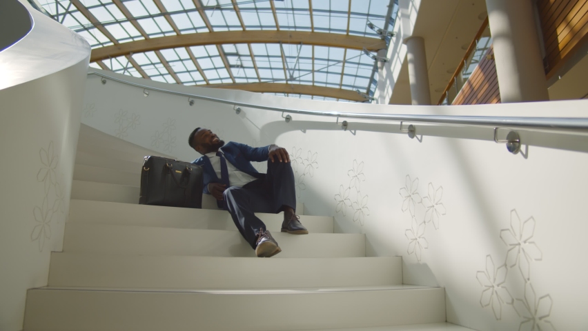 African man in formal suit and tie sitting on indoors office stair relaxing. Smiling young afro-american entrepreneur enjoying work break resting on staircase