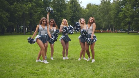 Group of excited cheerleading women shaking pompons smiling at camera. Portrait of young cheerleaders team in uniform posing at camera with pom poms in park