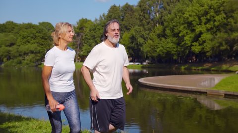 Happy caucasian old couple jogging running outdoors in park. Mature husband and wife doing cardio exercises running together by riverside in summer park