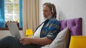 Modern senior man talk on video call on laptop lying in bed at home. Mature businessman having video conference using computer and headphones working remotely in bedroom