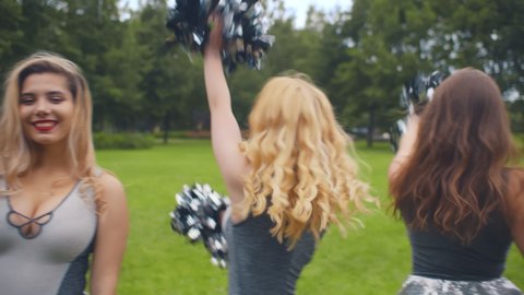 Portrait of dancing cheer team in uniform in summer park. Beautiful cheerleader women practicing outdoors with pompons turning around and smiling at camera