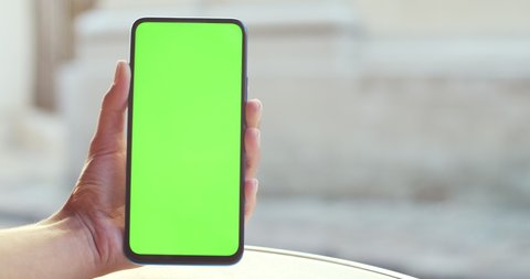 Close up view of female hand holding smartphone and pressing on mockup template screen. Concept of greenscreen and chroma key. People passing by at background