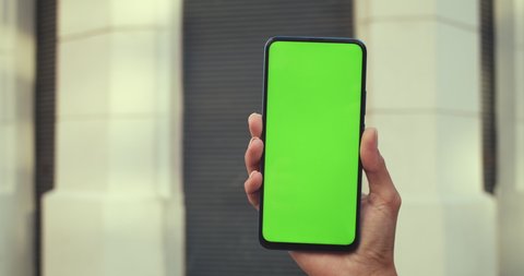 Close up view of female hand holding smartphone and pressing on green blank screen. Concept of mockup and chroma key. Closed building at background