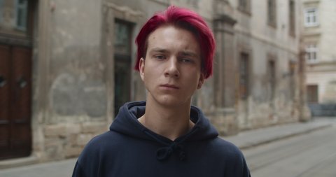 Crop view of depressed guy with dyed red hair looking to camera. Portrait of hipster millennial man with earring wearing dark hoodie standing at empty city street.Zoom in