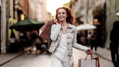 Young woman walks with colorful shopping bags on city street in slow motion. Outdoor portrait happy female tourist having good time wandering around town in summer