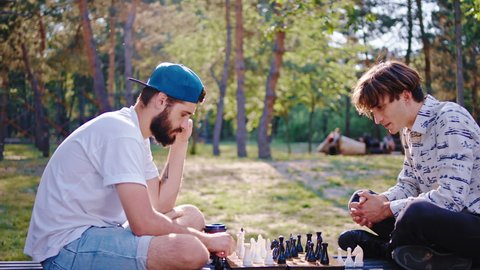 Two friends charismatic guys concentrated playing on a chess game in the middle of a large green park while sitting on the chair. 4k
