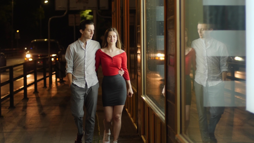 Young happy couple watch store showcase and see expensive thing they wish to buy behing glass frontage during night city walk