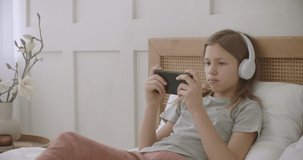 school girl is lying on bed in room and playing game on smartphone by internet, listening to music by headphones