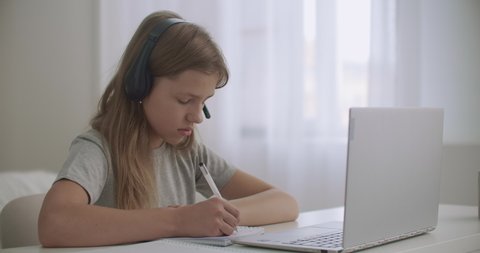 little girl is learning online by internet, listening teacher through headphones and looking at screen of notebook, writing notes in exercise book