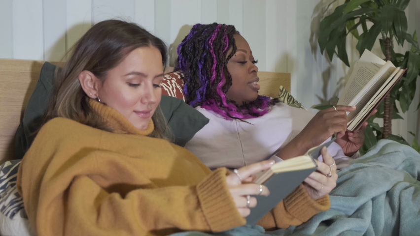 Close-up Portrait of African-American and Caucasian Woman Lying On Bed Reading Books, Home Furnishings, Cozy Apartment in the Evening, Ulybayutsya Laugh to Read the Study