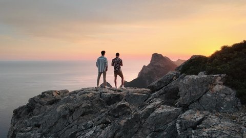 Aerial orbit shot of two men standing on a spectacular cliff and the sun rising on background
