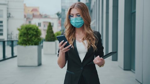Young business woman wearing protective face mask. Use mobile phone. Walking on street city near business center. Covid-19. Outdoors, coronavirus, corona, slow motion.