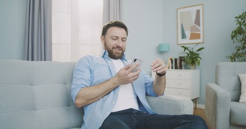 Middle plan of a handsome happy man at home, sitting down on a sofa while using smartphone, does various touching and swiping gestures. Man relaxing with mobile phone in cozy living room.