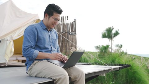 Young attractive happy asian guy using laptop computer work remotely anywhere using internet connected to office. Freelancer male work on vacation with scenic view of mountain, forest and tent resort.