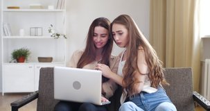 Two teenage girls sitting on couch looking at laptop screen, read online, watch videos or photos on network, hug spend time at home together. Sister comforts her friend stroking hair, friendly support