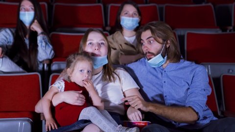 Family in masks in movie theater. Media. Young beautiful family watching cartoon in movie theater during pandemic. Family with child wearing masks in cinema