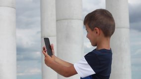 Cheerful boy is making a selfie at the phone against the background of beautiful white columns