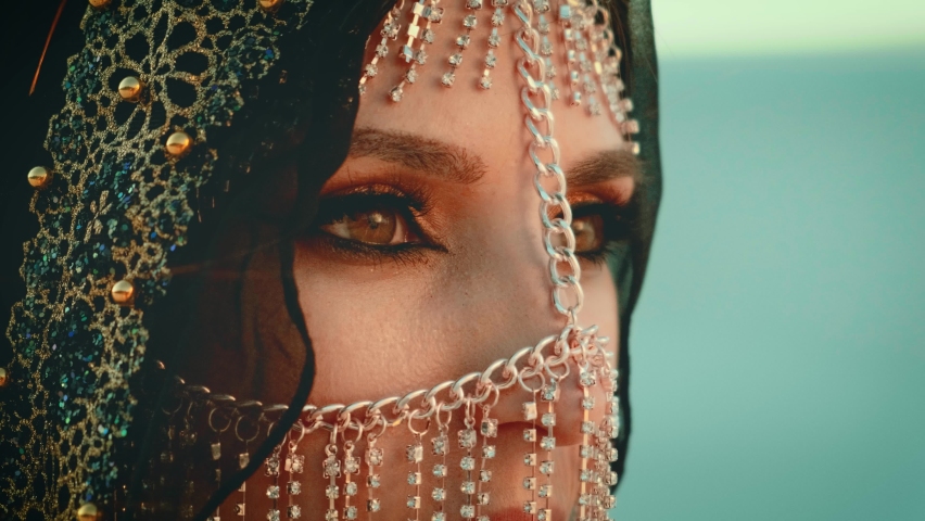 Close-up portrait young arabian woman. Beautiful brown eyes oriental makeup. Gold accessories metal chain golden mask hides face. Hijab shawl black color scarf covers head fashion style saudi arabia Royalty-Free Stock Footage #1059164603