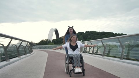 Excited teen daughter driving happy disabled mother on wheelchair along city bridge. Cheerful females having fun outdoors, joint leisure activity, positive lifestyle with disability, family bonding