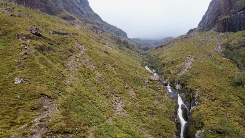 Flying low to the ground uphill the stream hidden in between the cloudy three Sisters mountains in Scotland