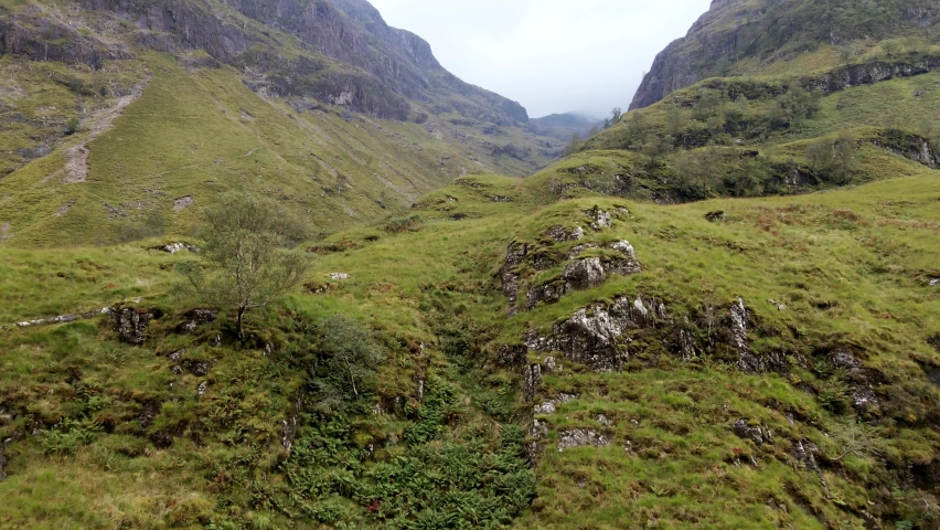 Flying low to the ground to reveal the stream hidden in the  cloudy canyon in between scotlands Three Sisters mountains
