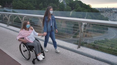 Handicapped woman on wheelchair and teen daughter in protective masks walking across city bridge. Mom with disability enjoying leisure together with teenage girl amid coronavirus infection outbreak