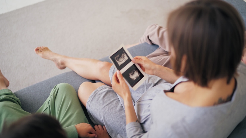 Beautiful pregnant woman and her sons are holding ultrasound scan on her belly in sofa at home, pregnant and family concept.