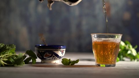 Slow motion wide shot of hand picking up an ornate teapot and pours tea into a glass with Moroccan bowl and mint around.