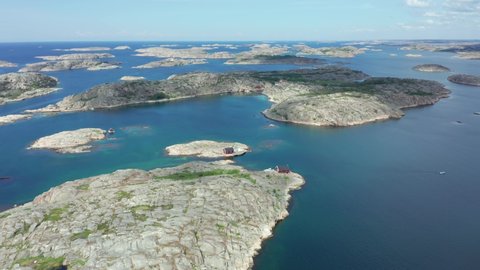 Sweden west coast in aerial drone shot. Swedish coastline with small rocky islands or smooth cliffs with red fishing sheds and tiny wooden cabin outhouses on a sunny day. Clean blue ocean landscape