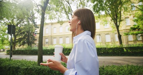 Authentic shot of young happy smiling business woman is making a conference call while drinking a morning coffee and taking a relaxing walk in a green city park by the way to an office in a sunny day.