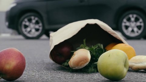 A paper bag of food falls to the pavement. Apples roll out of the bag, the baguette breaks. Bread crumbs fly when they hit the ground. Grocery bag falling on the street. Slow motion. Close up.