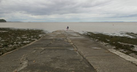 Grandparent and grandchild explore coastal slipway edge watching the sea on cloudy day, wide shot