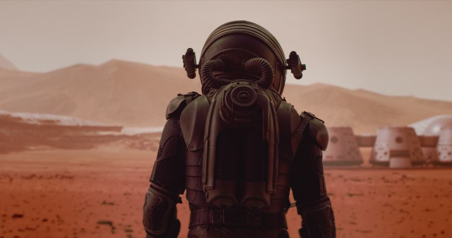 HANDHELD TRACKING back view of astronaut wearing space suit walking on a surface of a red planet. Martian base and rover in the background. Mars colonization concept Royalty-Free Stock Footage #1059183989