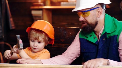 Father and son in protective helmets hammer a nail into a board. Dad teaches his boy a building