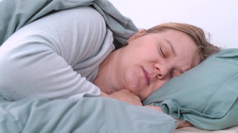 Young blonde tired woman in pajamas is sleeping, mumbling and snoring in bed under warm blanket at home. Health and breathing problems, somnambulism
