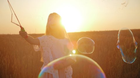 Little girl blowing soap bubbles in wheat field at sunset time. Slow motion video.