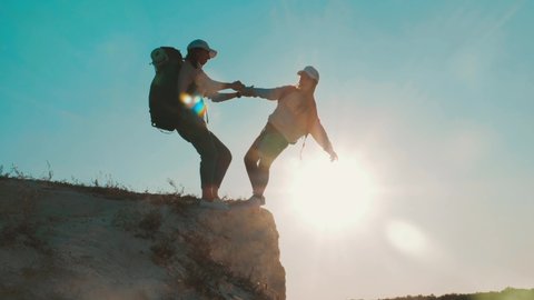 Girl helps her friend climb up the last section of mountain. Tourists with backpacks help each other, silhouette with sunset. The joint work teamwork. Tourism, travel and healthy lifestyle concept.