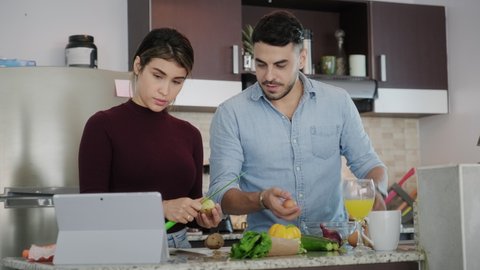 Young couple cooking food together at home, watching tutorial video for recipe on tablet. Happy man and woman preparing omelette. Husband and wife smiling in kitchen. Married partners and lifestyle