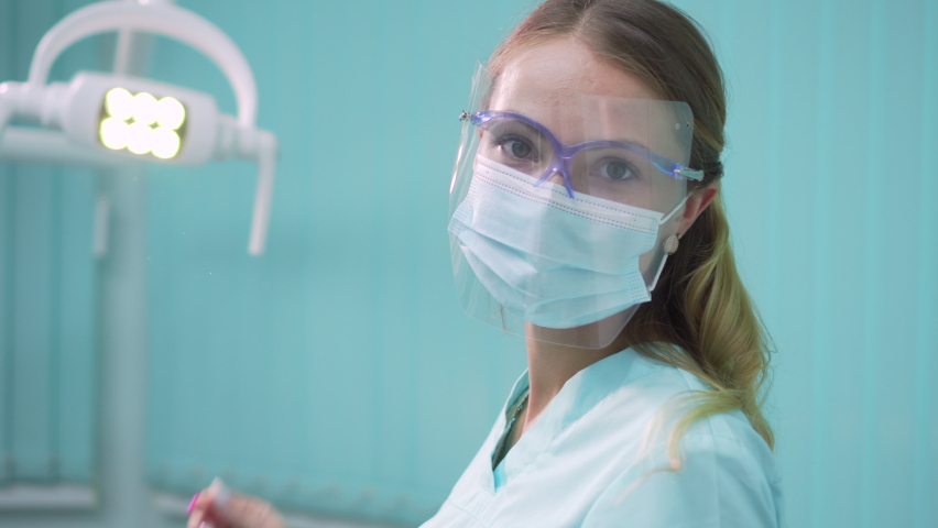 Young pretty woman dentist puts on a protective face mask and screen and then looks at the camera. Close-up. | Shutterstock HD Video #1059190004