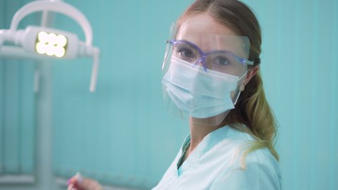 Young pretty woman dentist puts on a protective face mask and screen and then looks at the camera. Close-up.