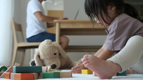 Asian preschool toddler girl playing with building blocks at home. Father in white T-Shirt working on the wooden table. Stay home. Quarantine remote work