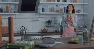 Woman in apron drinking coffee near ingredients in kitchen